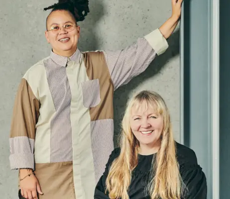 Kaksi naista istuu valokuvaa varten - yksi blondi istuu mustalla päällään ja toinen seisoo raidallinen nappi päällä