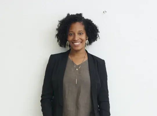 Nainen bleiserissä ja harmaassa paidassa seisoo valkoisen seinän edessä