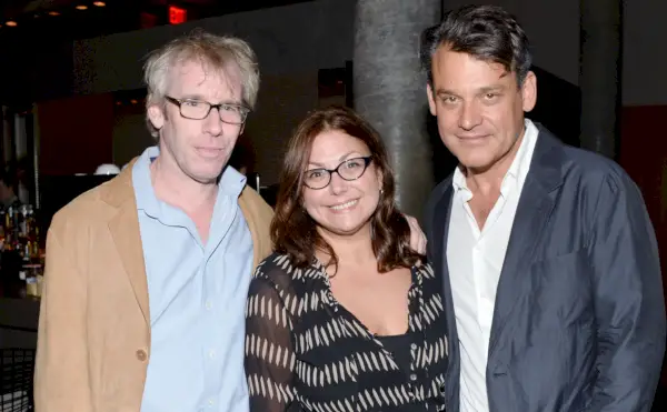 Tre personer poserer til foto, to mænd i jakkesæt flankerer en kvinde i kjole.