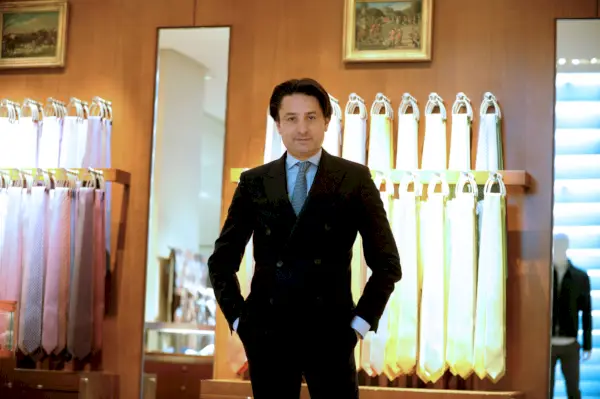 Mężczyzna w średnim wieku z czarnymi włosami pozuje w czarnym garniturze przed wieszakiem z krawatami.