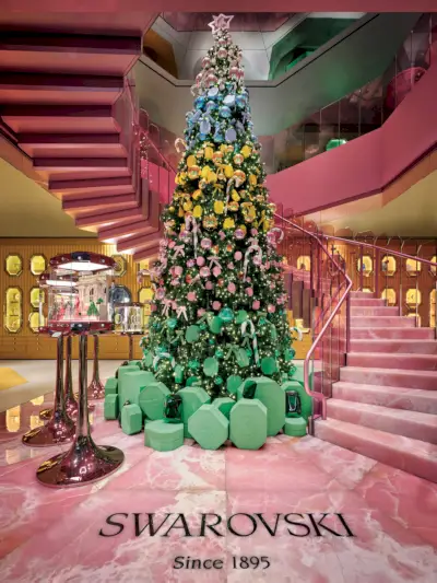 Lahjalaatikoihin ja monivärisiin koruihin koristeltu korkea joulukuusi kauppatilassa, jossa on kiemurteleva vaaleanpunainen portaikko