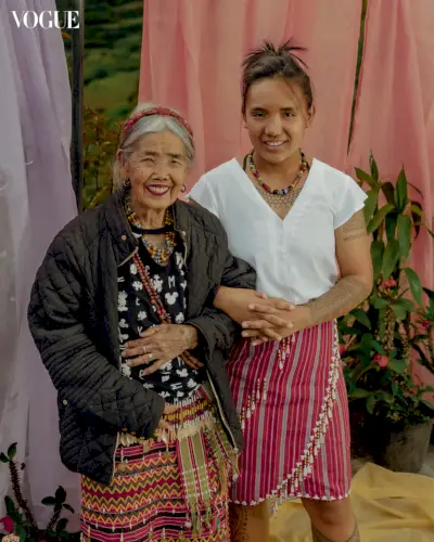 Vanhempi nainen ja nuorempi tatuoitu nainen seisovat kädet yhdistettyinä kangastaustan edessä