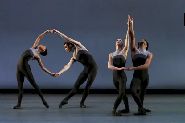 Neljä tanssijaa musta-harmaissa puvuissa poseeraa paljaalla lavalla.