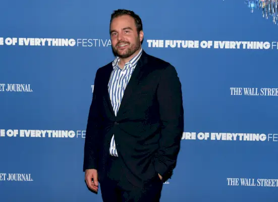 Charles Forelle hymyilee puku yllään sinisen kyltin edessä.