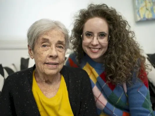 Nainen, jolla on pitkät kiharat hiukset ja silmälasit, nojaa ikääntyneen lyhyen harmaatukkaisen naisen olkapäälle