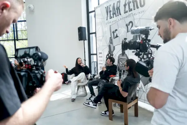 Una tavola rotonda con tre uomini, uno dei quali parla al microfono e gesticola, davanti alla grande copertina di un album in bianco e nero mentre un fotografo e un operatore video guardano