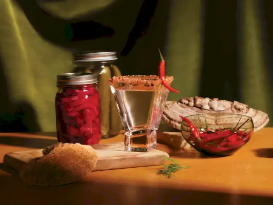 Asetelma suolakurkkupurkkeja, paprikakulhoja, punasipulipurkkeja, mintun oksia ja puukaiverruksia, jotka ympäröivät hienoa suolakurkku- ja pippurimartinia puulaudalla.