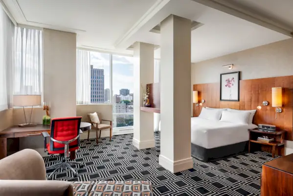 En suite på Sofitel Montreal med et siddeområde til venstre, søjler foran en seng til højre og udsigt over byen i baggrunden.