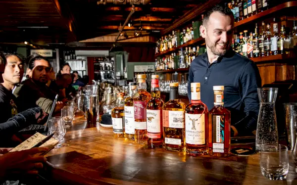 Persone sedute a un bancone di legno con dietro un barista sorridente, parzialmente nascosto da una fila di bottiglie di whisky.