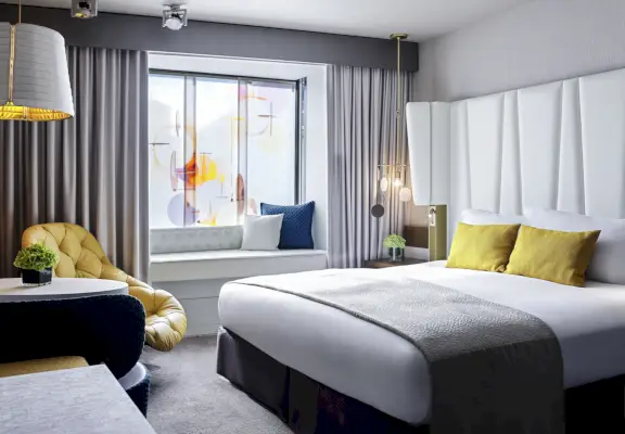 Pokój gościnny w hotelu Sofitel Monachium Bayerpost z łóżkiem po prawej stronie, lampą i krzesłem po lewej stronie oraz oknem w tle.