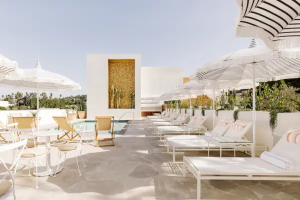 Basen na eleganckim patio z białymi parasolami i leżakami z brązowymi i różowymi akcentami, otoczony roślinami w białych donicach.