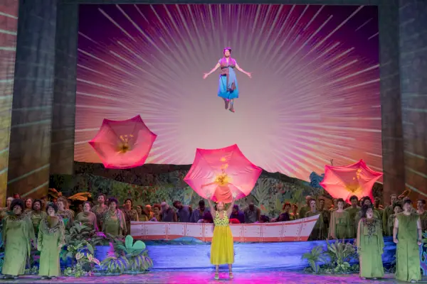 Sinipukuinen nainen kelluu kuoron jäsenten lavan yläpuolella värikkäässä oopperassa